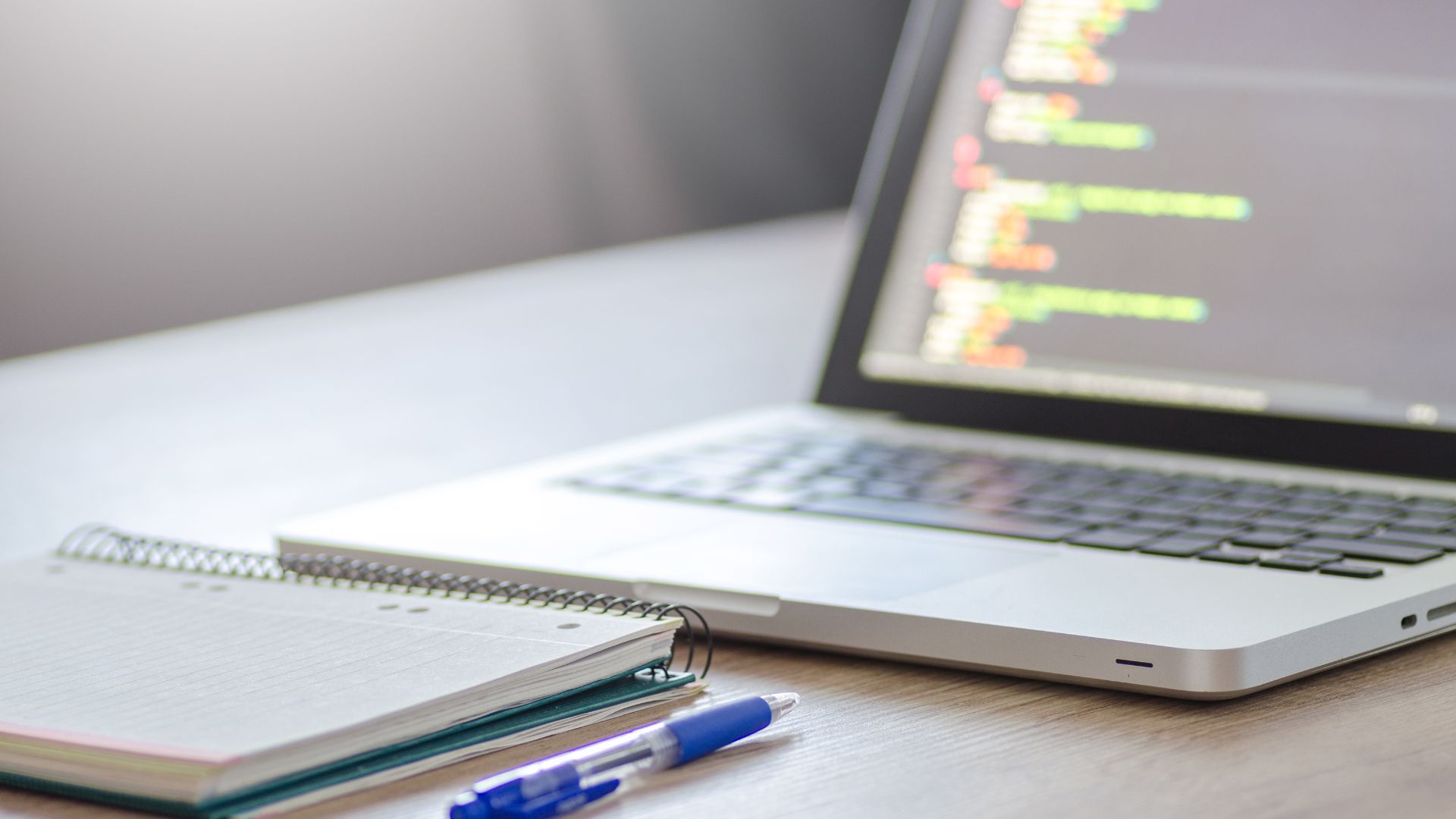Laptop displaying code next to a notepad and pen on a desk.