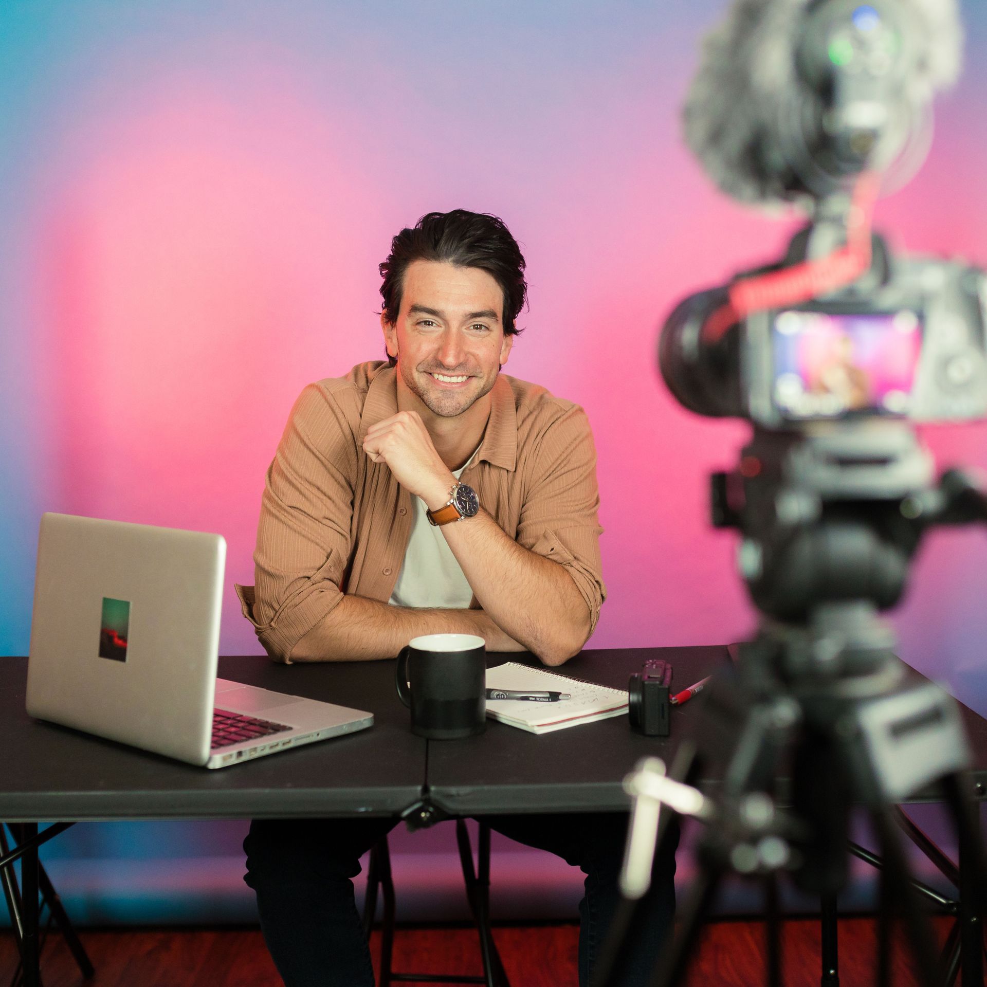Rollers Media: Man at desk with laptop and coffee cup smiling at camera, studio with pink and blue backdrop.