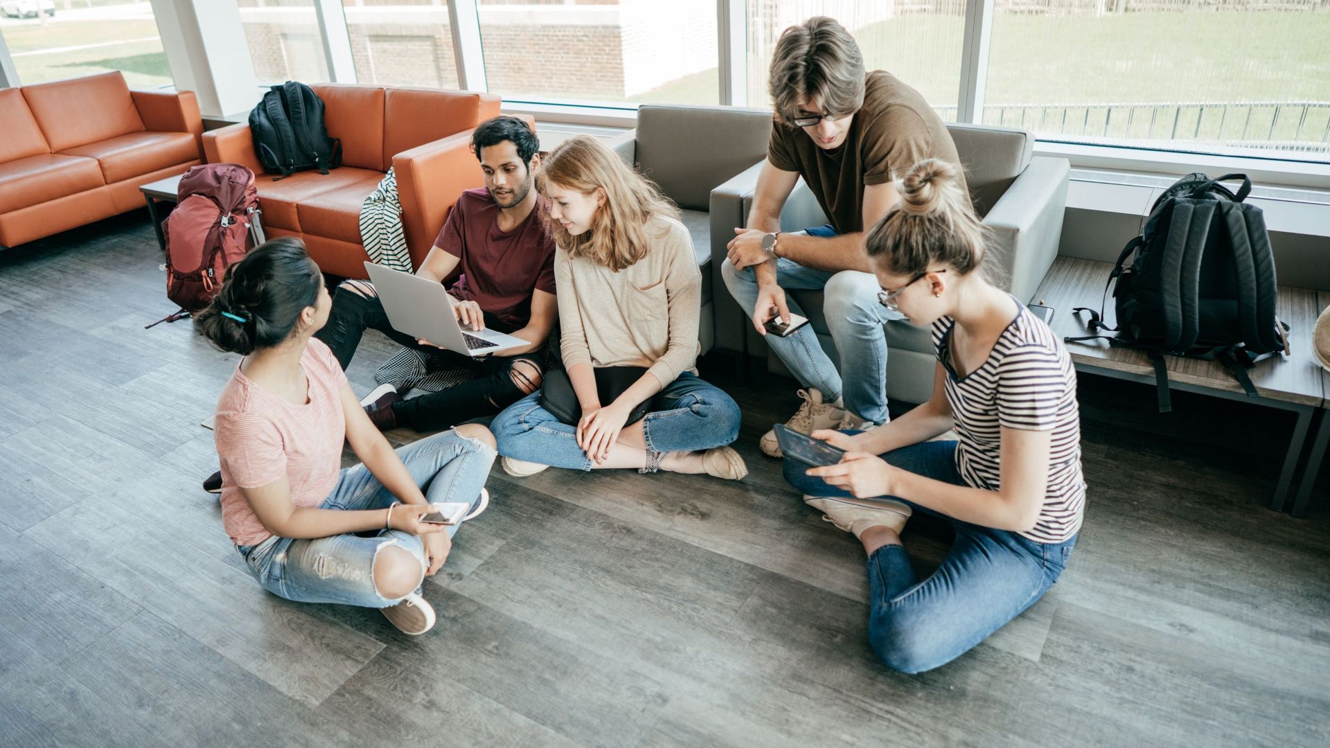 Group of students collaborating on the floor, using a laptop and devices in a brightly lit lounge.