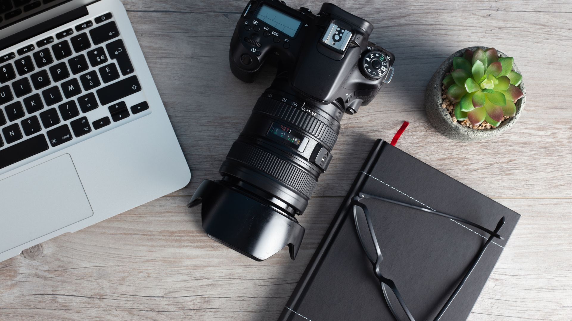 Laptop, camera with lens, notebook, glasses, and plant on a wood desk.