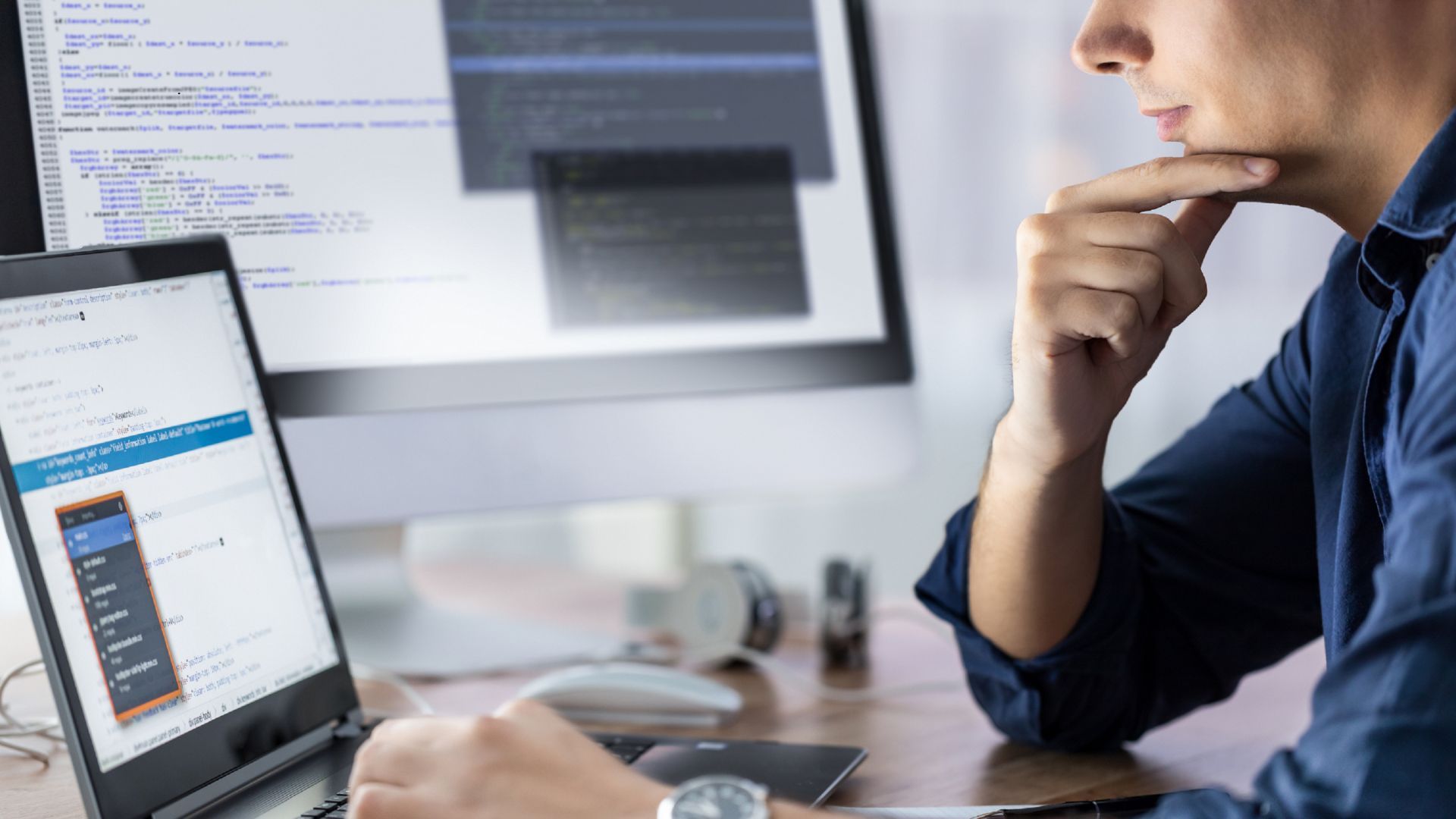 Man in blue shirt, pondering, looks at code on a laptop and monitor in an office setting.