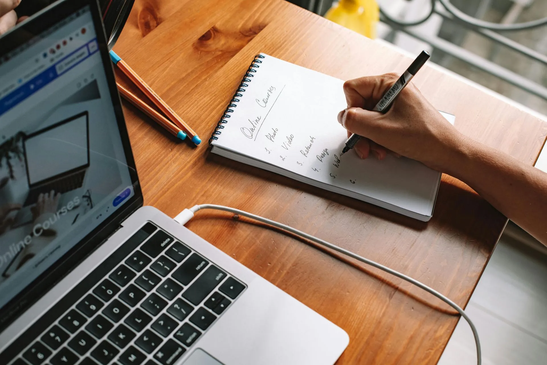 Laptop and notepad on a wooden desk; hand writing on the notepad.