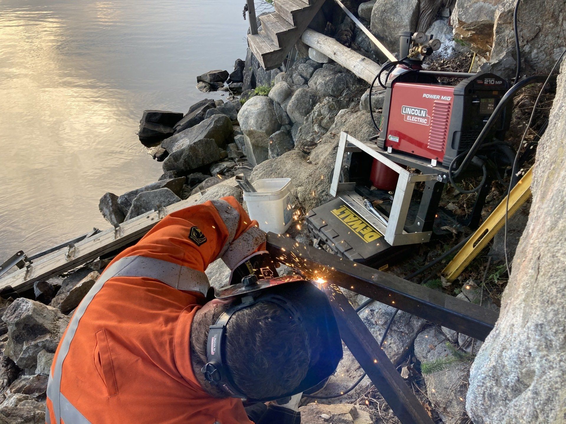 A man is welding a piece of metal next to a body of water.