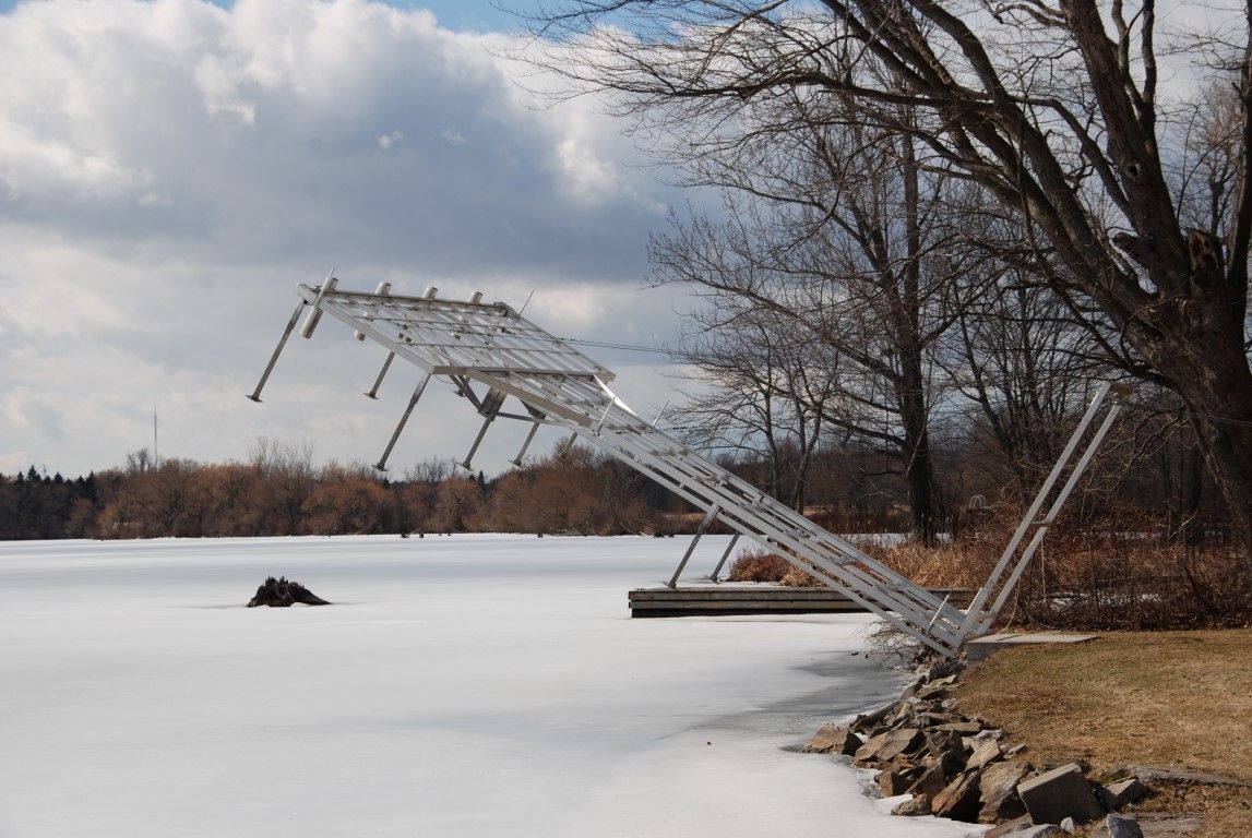 Boat lift frame tipped and leaning over frozen lake shoreline after winter ice damage in Ontario.