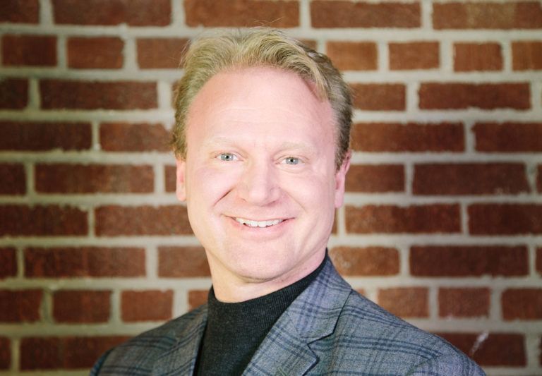 A man in a suit is smiling in front of a brick wall.