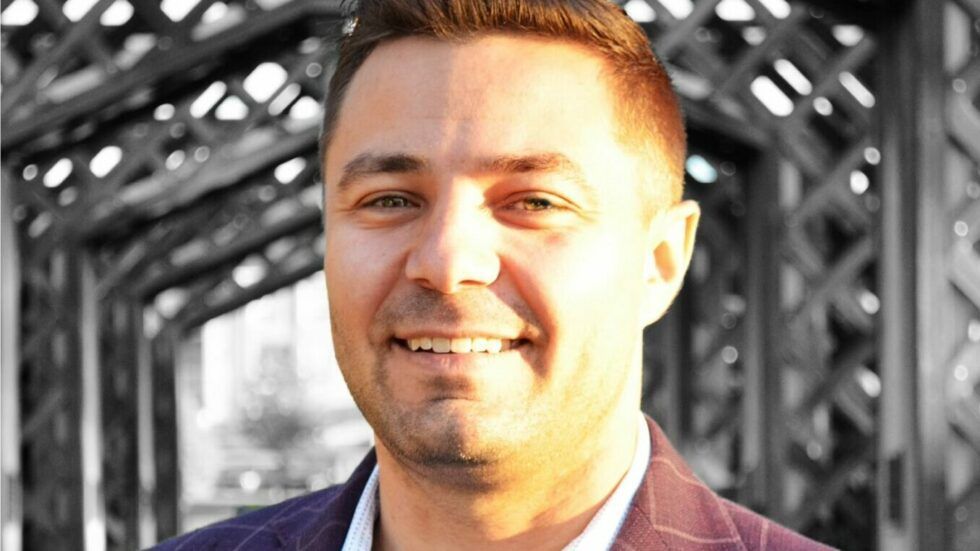 A man in a suit is smiling in front of a bridge.