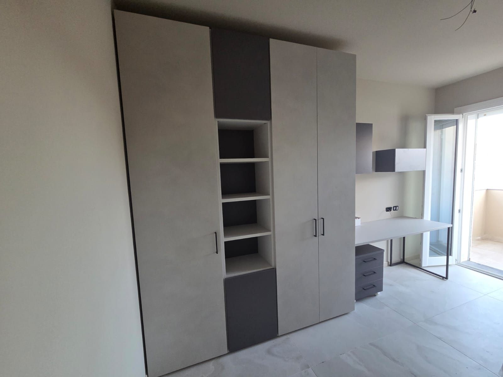 Un armadio a muro con ante grigie e bianche, ripiani aperti e una scrivania con una porta-finestra sullo sfondo.