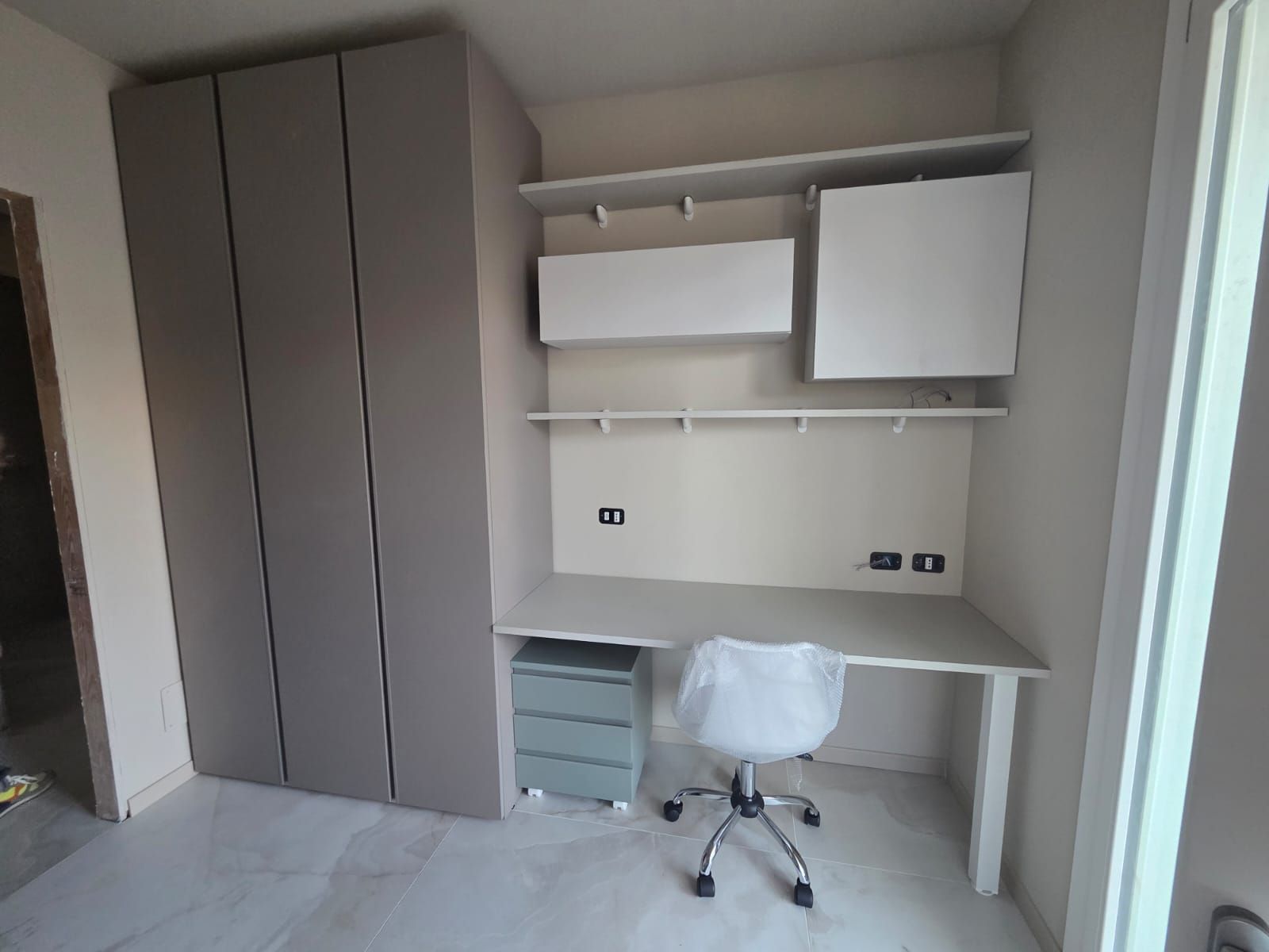 Scrivania integrata con armadietti, scaffali e una sedia in una stanza. Colori neutri. Armadio a sinistra.