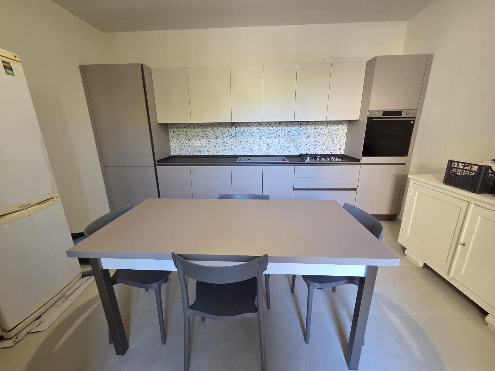 Cucina con mobili chiari, ripiani grigi, tavolo da pranzo, sedie ed elettrodomestici.