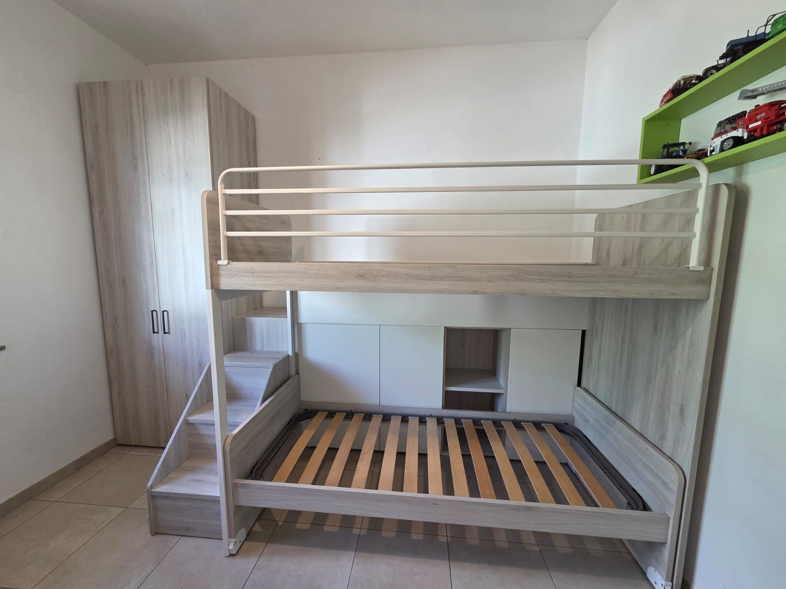 Letto a castello con gradini, un armadio e uno scaffale con macchinine giocattolo contro una parete bianca.