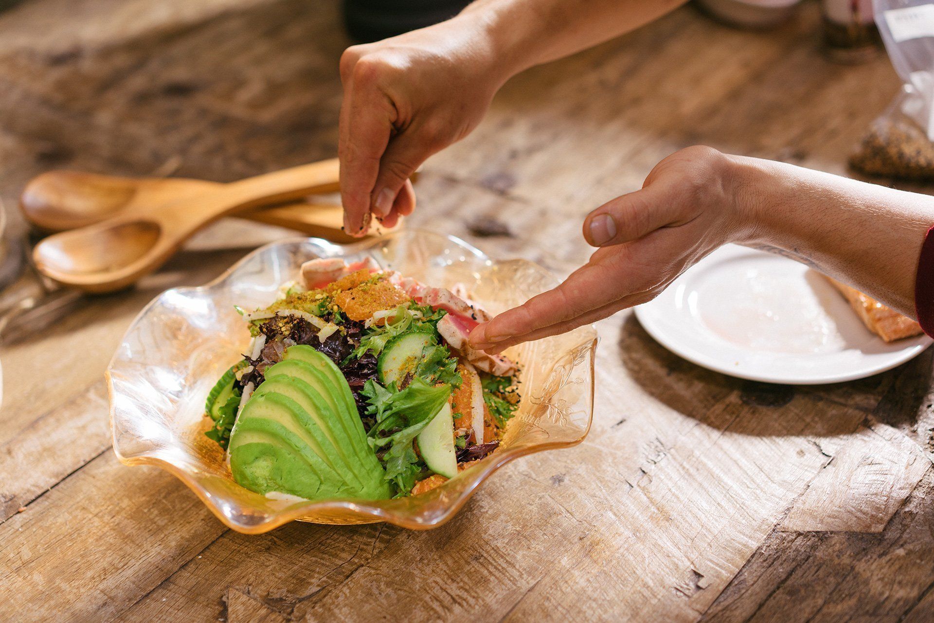 A person is adding avocado to a salad on a wooden table.