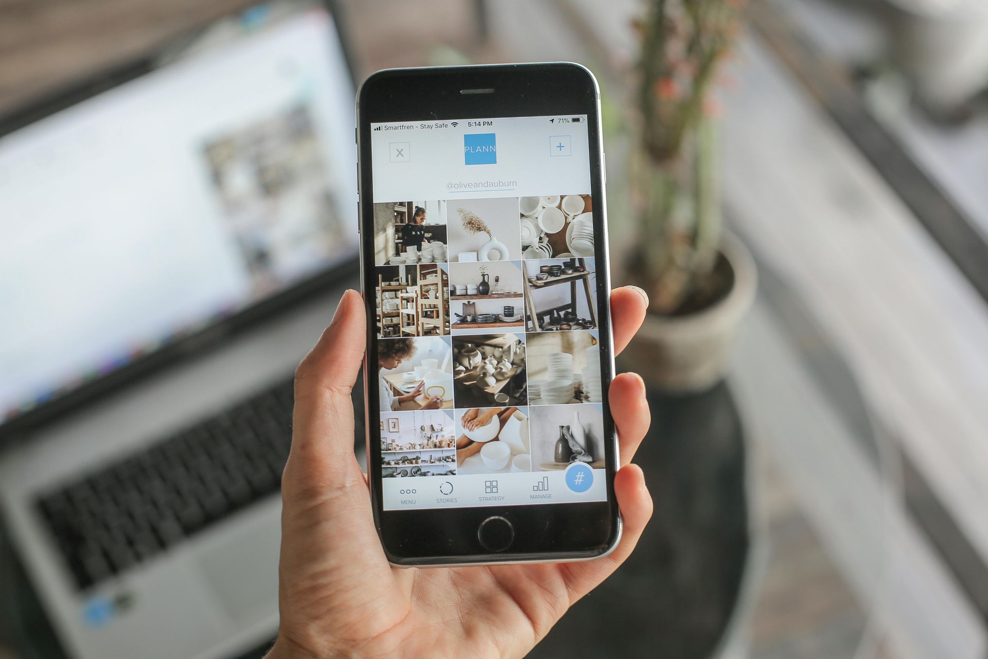 Hand holding a smartphone showing a photo gallery, with a laptop blurred in the background