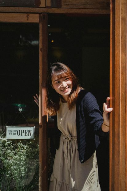 Smiling woman in a beige dress stands in a doorway, holding the door open under a small OPEN sign.