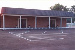 A red brick commercial building with a dark metal awning and white support columns above an asphalt parking lot.
