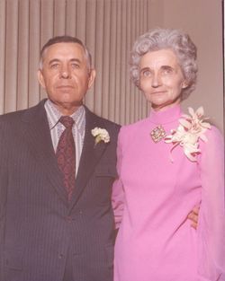 A person in a dark suit and tie standing beside another in a pink long-sleeved dress with a floral corsage.