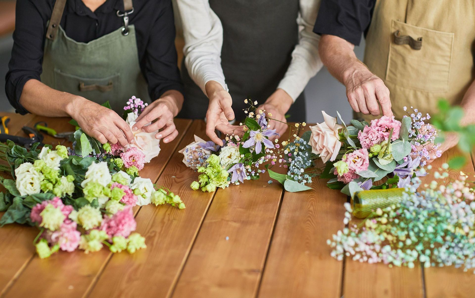 Three people wearing aprons arrange colorful bouquets of flowers on a wooden table.