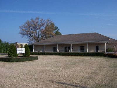 A beige funeral home building with a large brown roof, a covered porch, and a lawn sign under a bare tree against a blue sky.