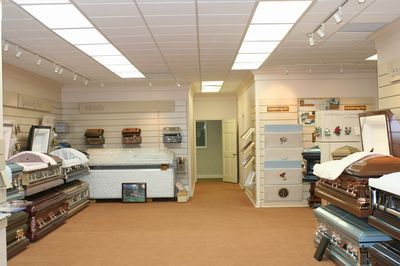 A showroom displaying various caskets, with a neutral-toned interior, lighting tracks, and a hallway in the background.