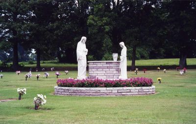 Statues of two figures stand by a central stone well, surrounded by pink flowers in a cemetery with trees in the background.