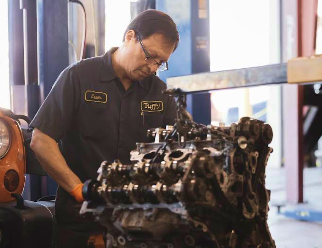 Mechanic examining a car engine in a garage. He is wearing glasses, dark clothes and is focused on the engine.