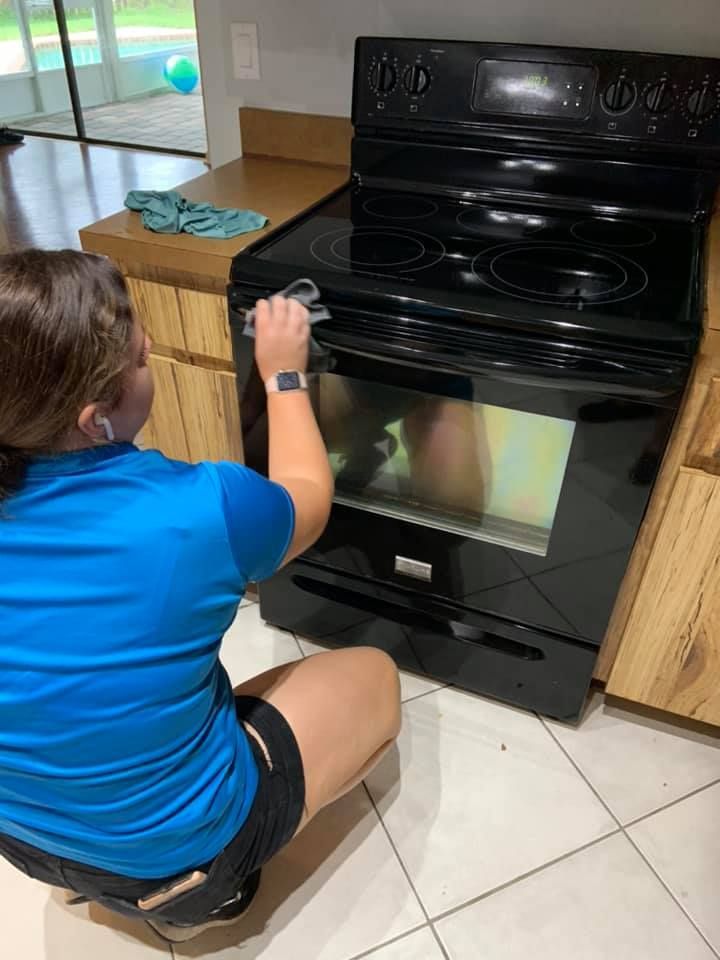 A person lies on a wooden floor, reaching under a stainless steel dishwasher to adjust the kickplate.