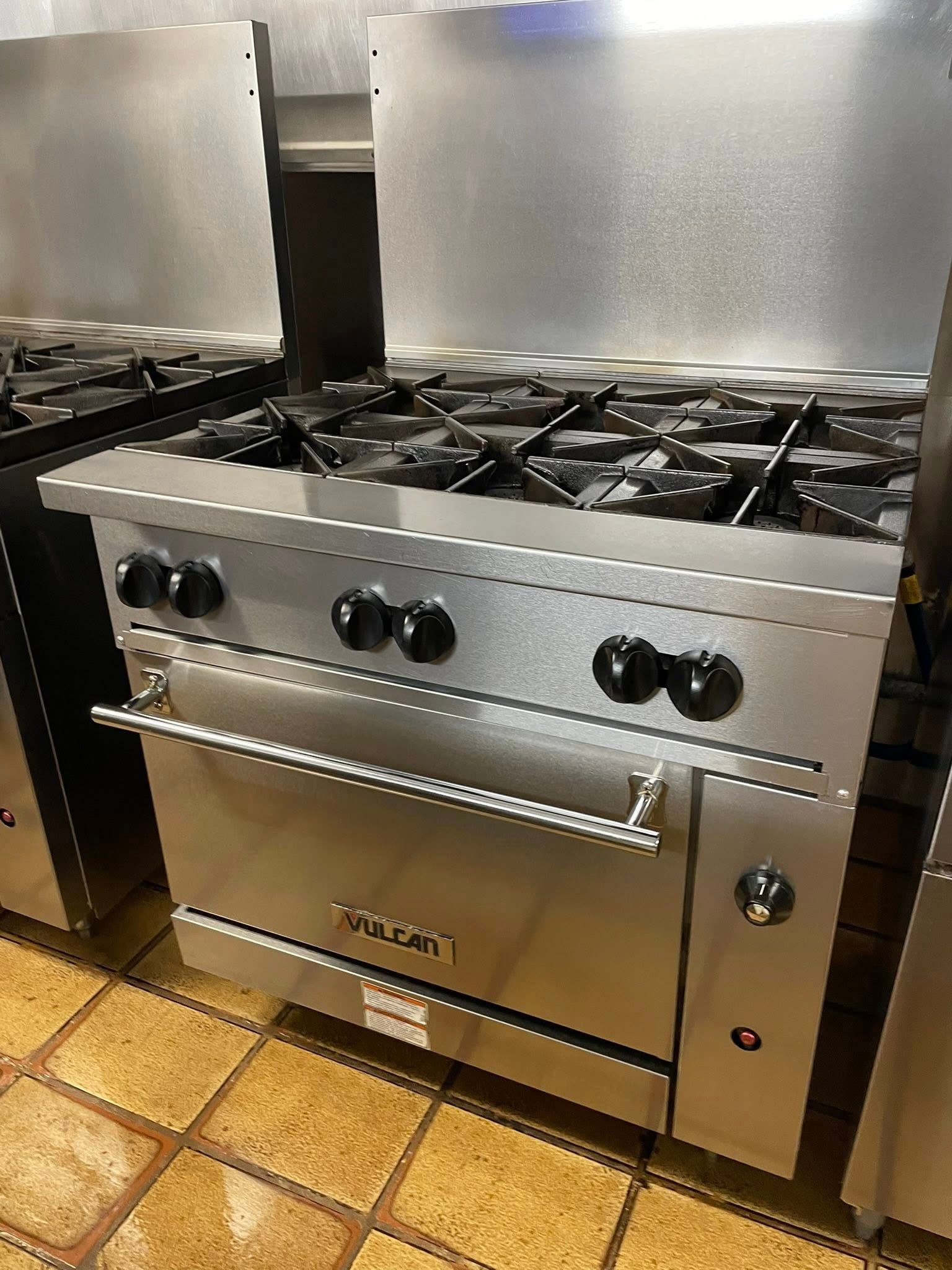 Stainless steel commercial gas range oven in a kitchen, with multiple burners and black control knobs.