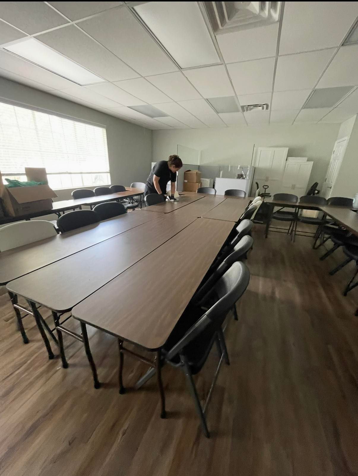 Conference room with long table, black chairs, large windows, and two people near the back wall.