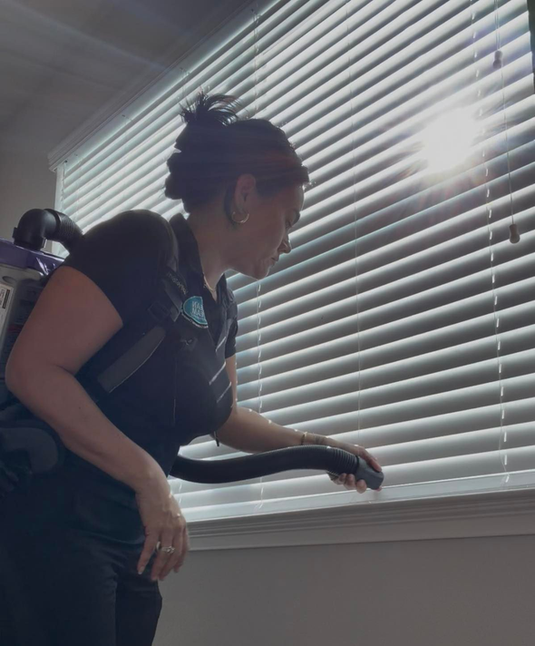 Person in dark clothing peering out a window with white blinds, sunlight streaming in.