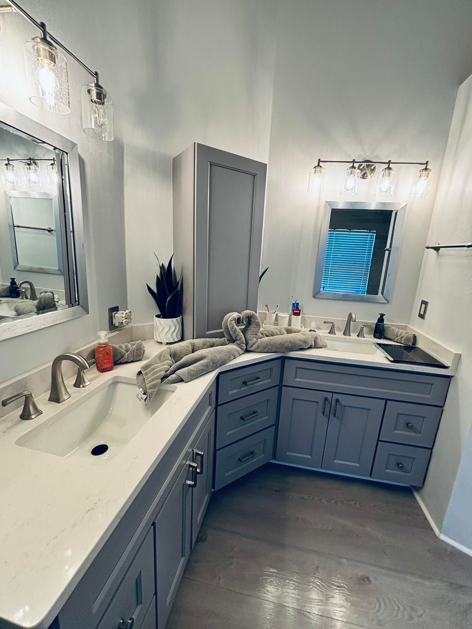 Bright bathroom with gray cabinets, large mirror, white counters, and a corner bathtub near a window.