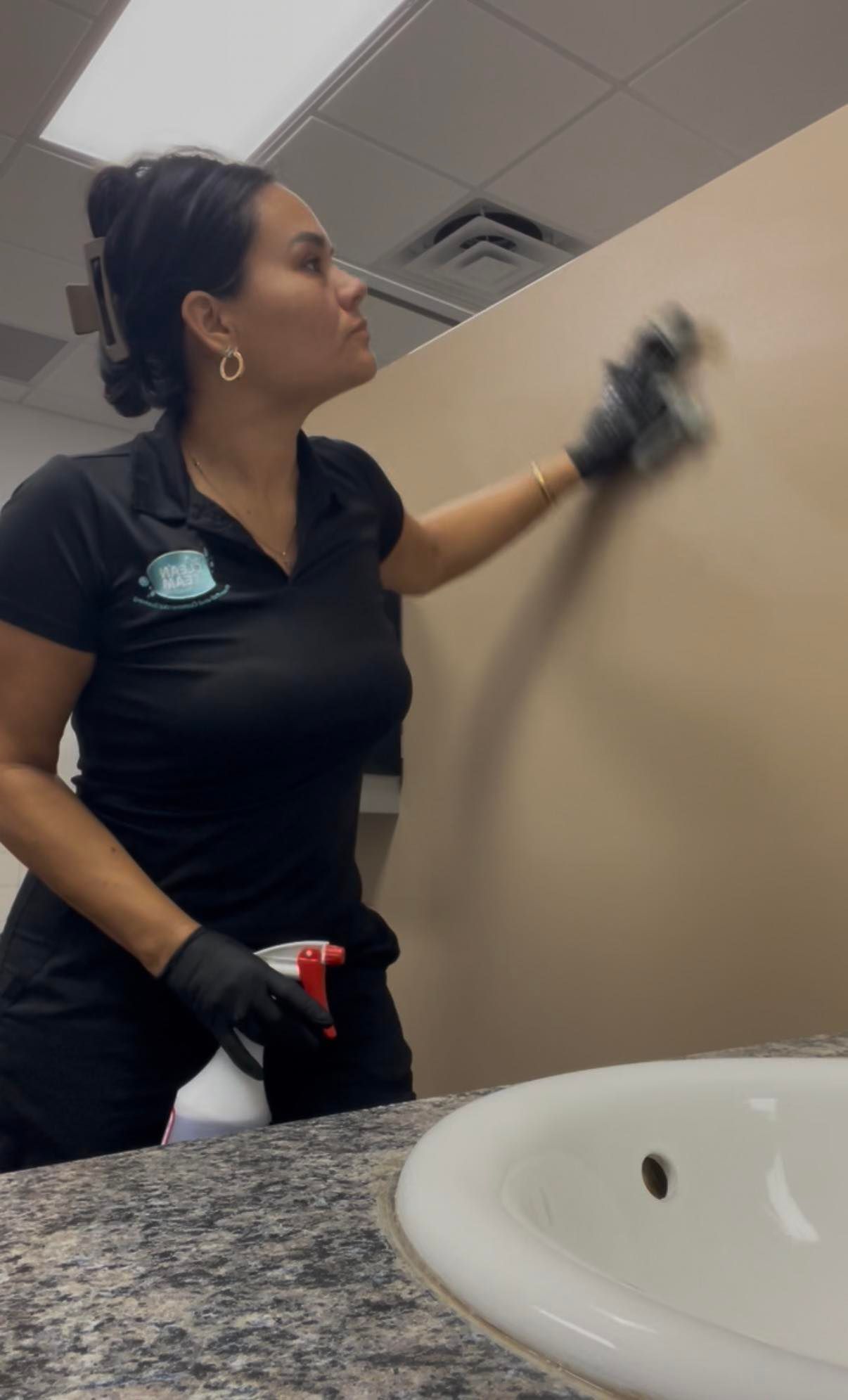 Woman cleaning a restroom mirror, wearing black gloves and a black shirt, near a sink and countertop.