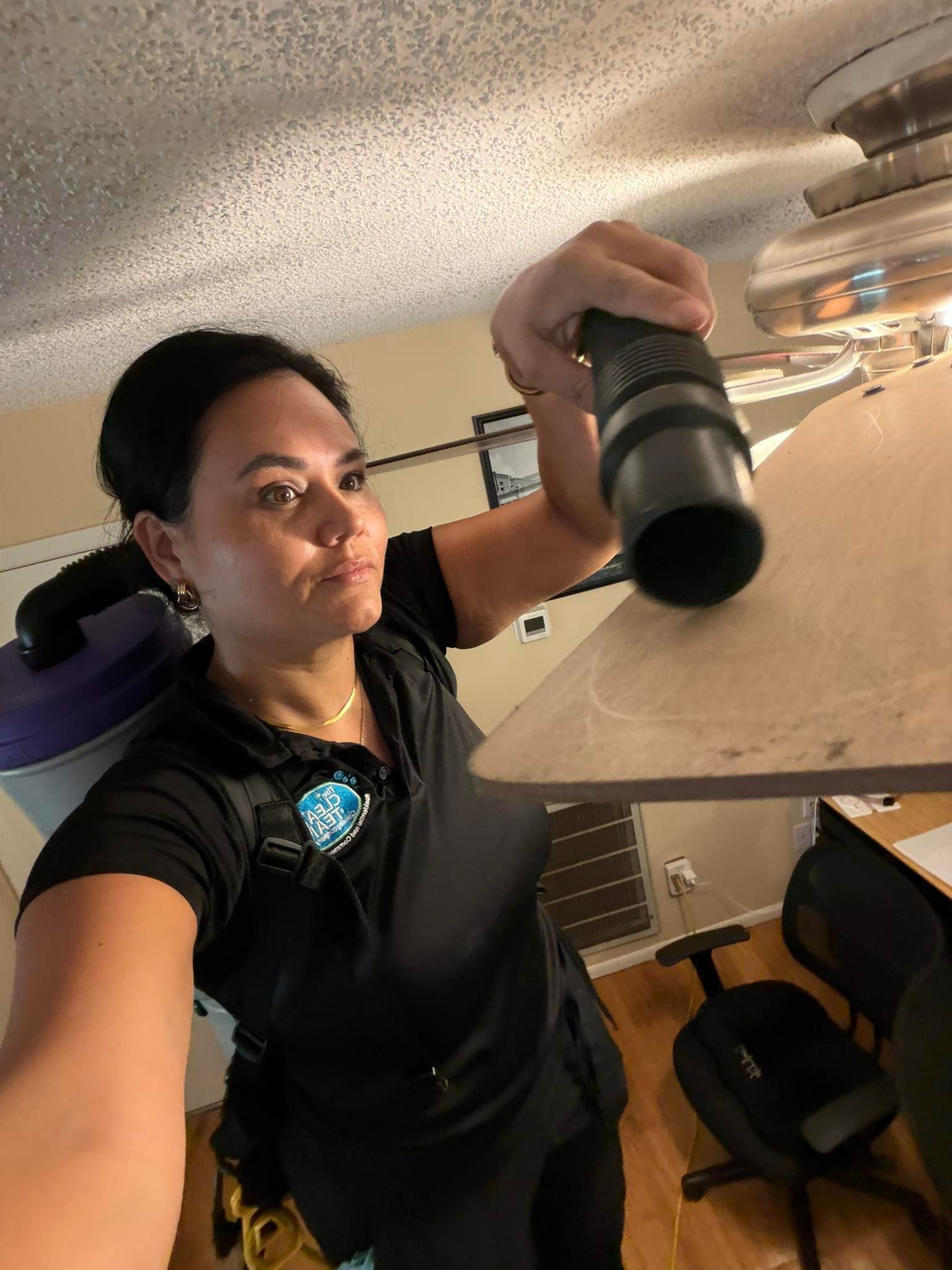 Person in black shirt taking a selfie in a room with a desk and chair, holding a camera