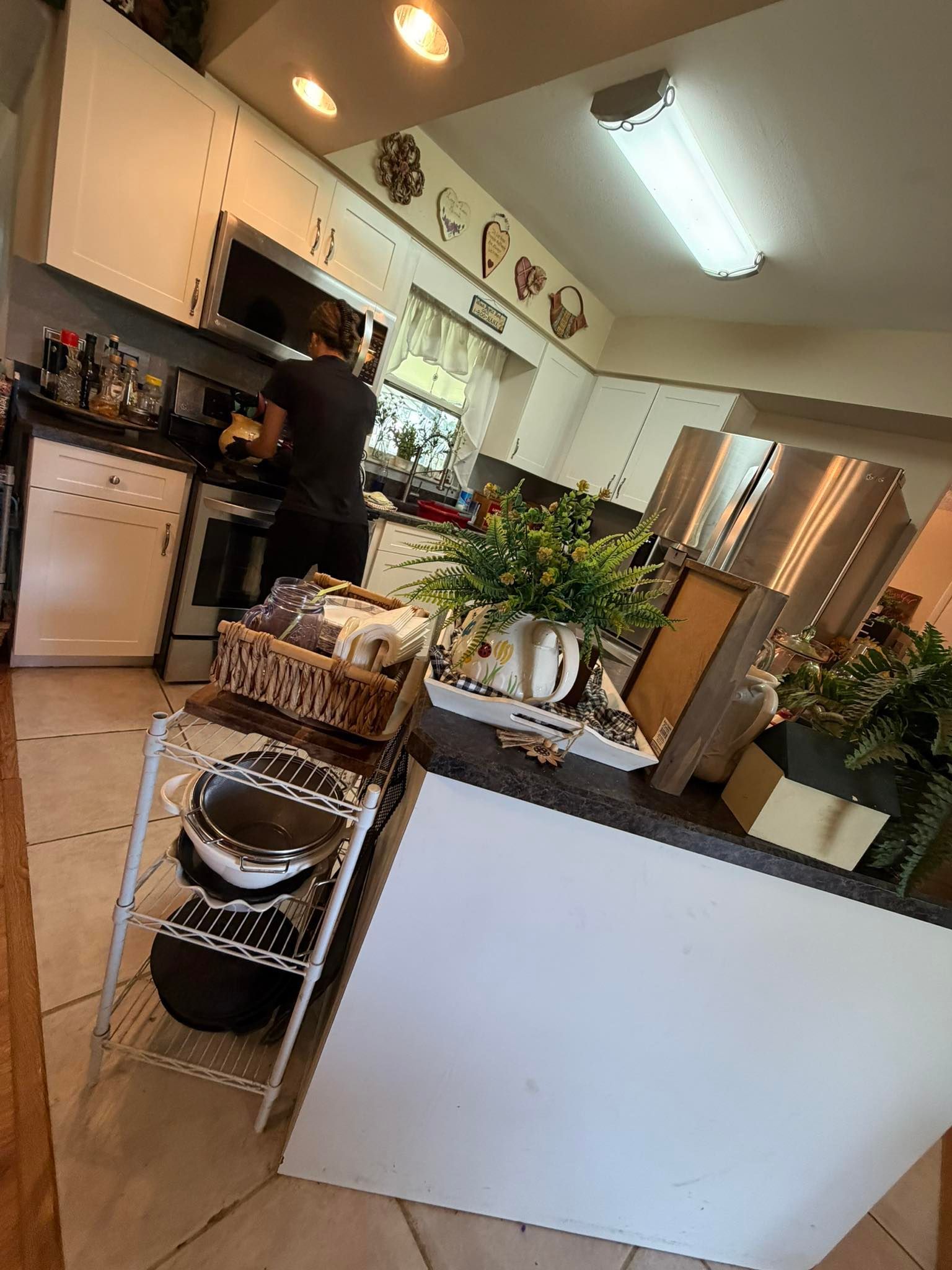 Bright kitchen with white cabinets, hanging lights, potted plants, and a person cooking near the stove.