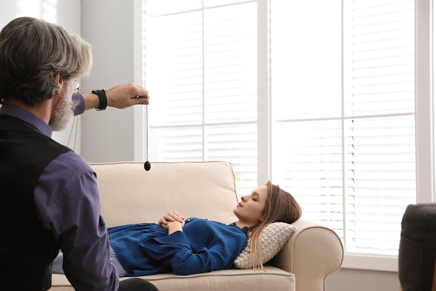 A woman is laying on a couch while a man holds a pendulum.