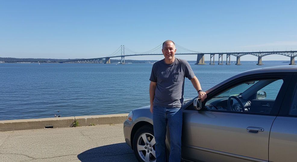 Smiling man standing confidently next to his car on waterfront by Chesapeake Bay Bridge, clear blue sky