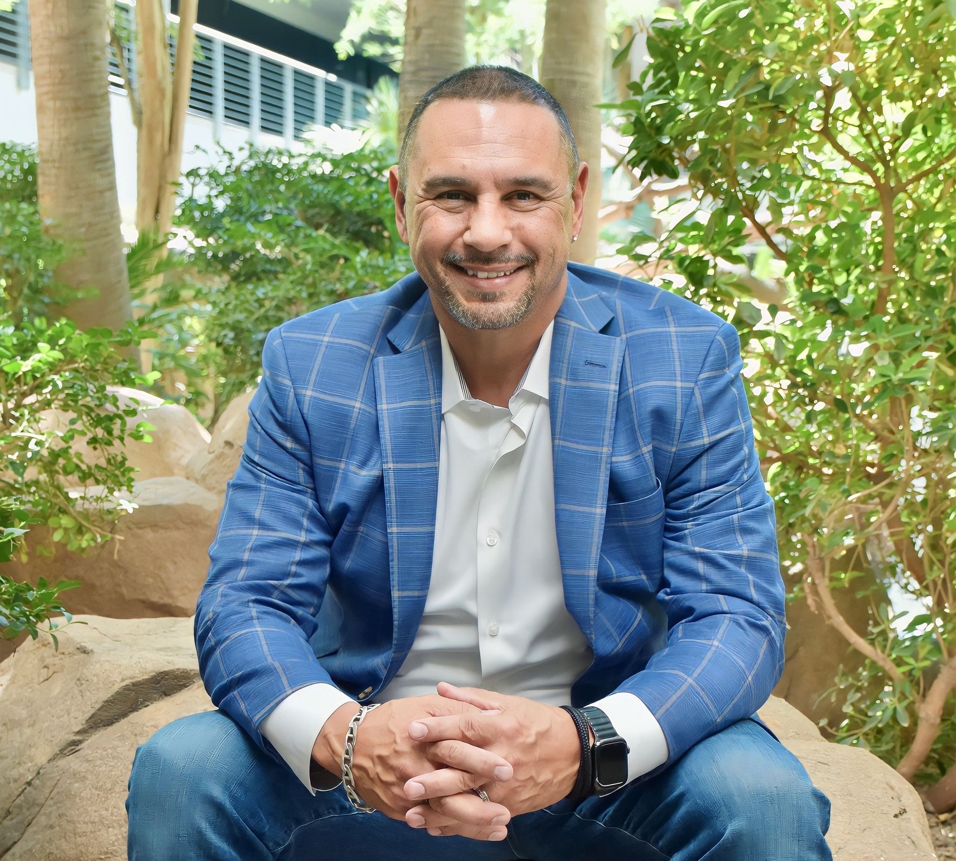 A man in a blue suit is sitting on a rock with his hands folded and smiling.