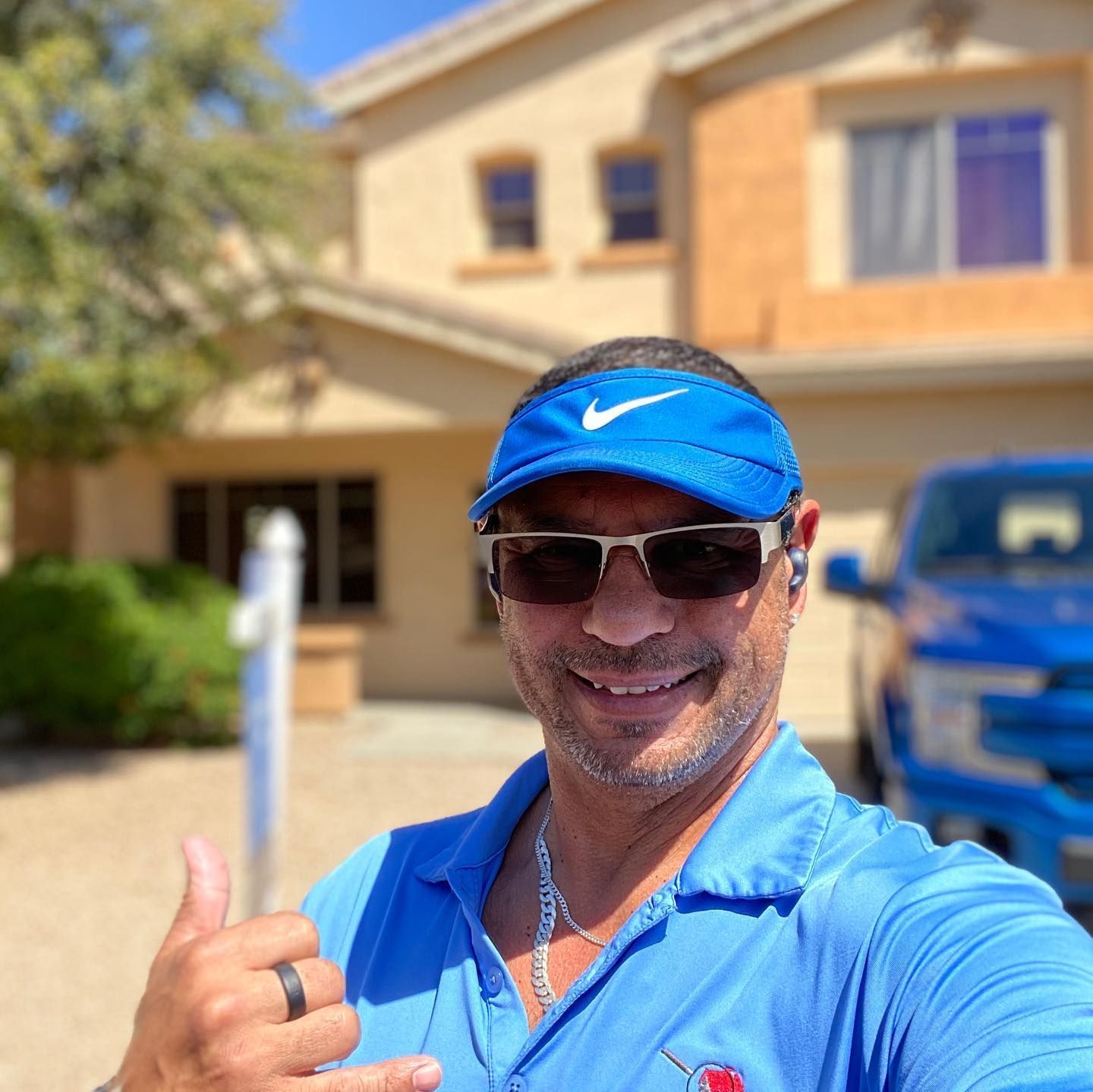 A man wearing sunglasses and a blue hat is giving a thumbs up in front of a house.