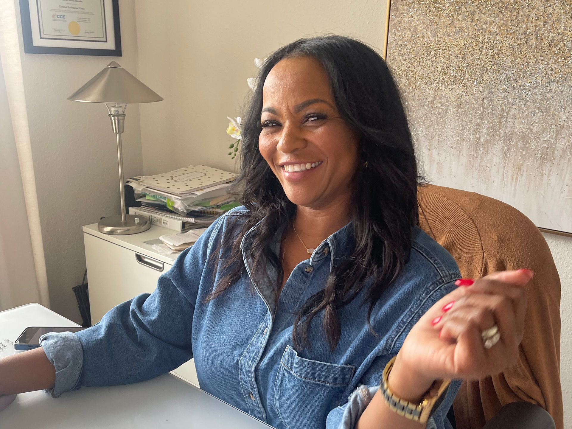 A woman in a red shirt is sitting at a desk with a computer and a tablet.