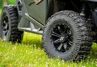 Black UTV tires on black rims in green grass with white flowers. | Impact Automotive