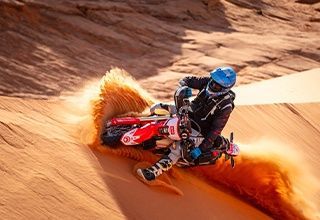 Motorcyclist leaning into a dune, kicking up sand. Red and white bike on a sandy slope. | Impact Automotive