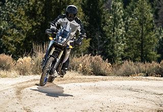 Motorcyclist riding a gray and black dual-sport motorcycle on a dirt road in a forested setting. | Impact Automotive