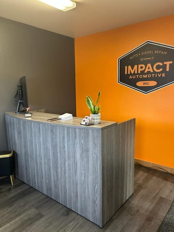 Reception area of Impact Automotive, with gray wood-look desk, orange and gray walls, logo, and a small plant. | Impact Automotive