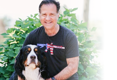Man with a dog, both looking at the camera. Man wears a shirt with an American flag rifle design. Green foliage in background. | Impact Automotive