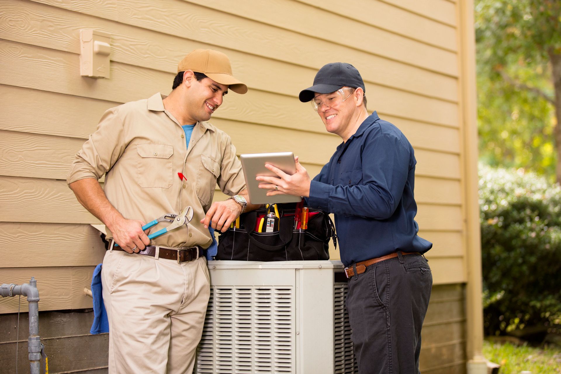 Two HVAC technicians reviewing data on a tablet next to an air conditioning unit outdoors.