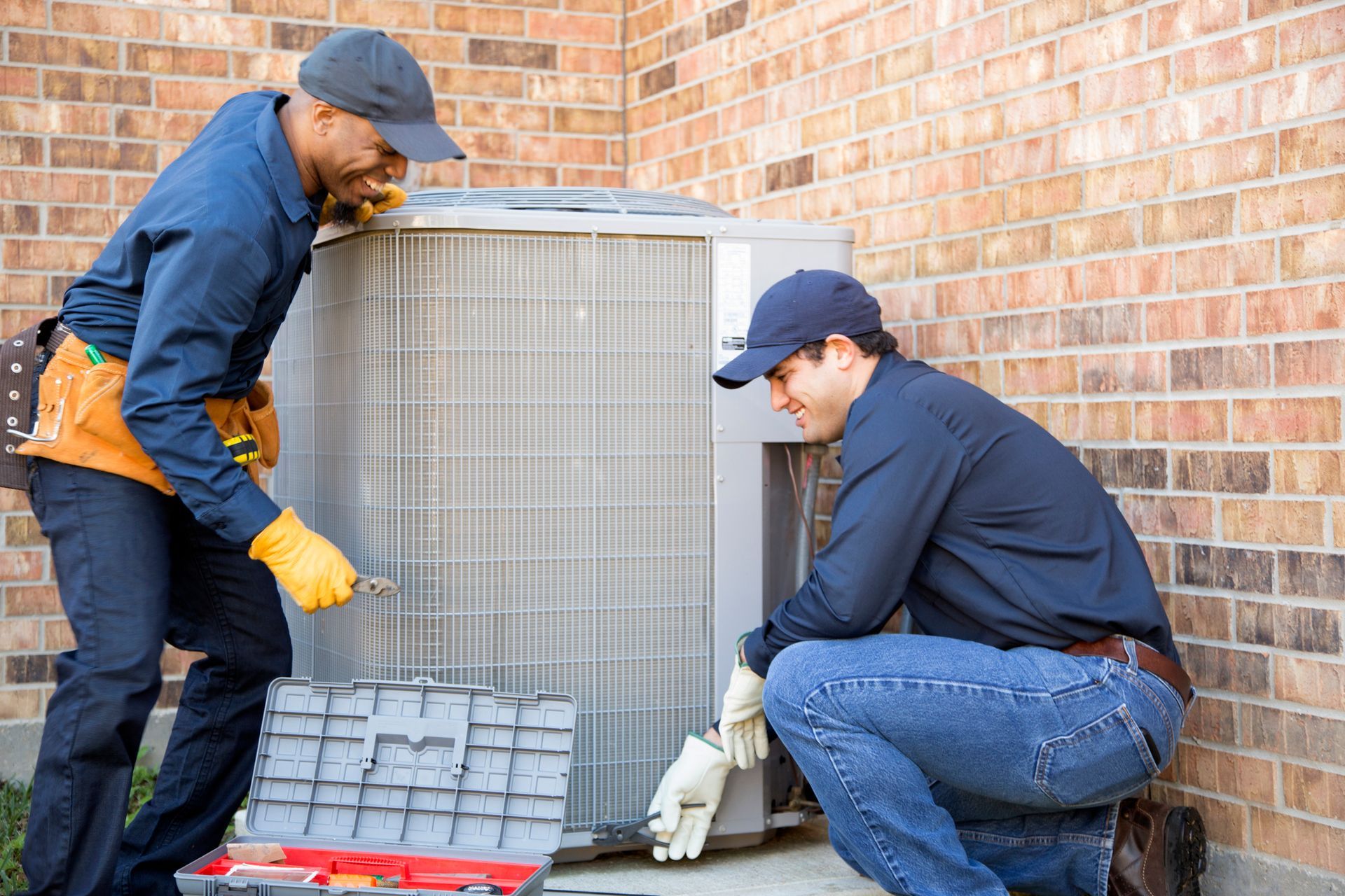 Two HVAC technicians working on an air conditioning unit outside a brick building.