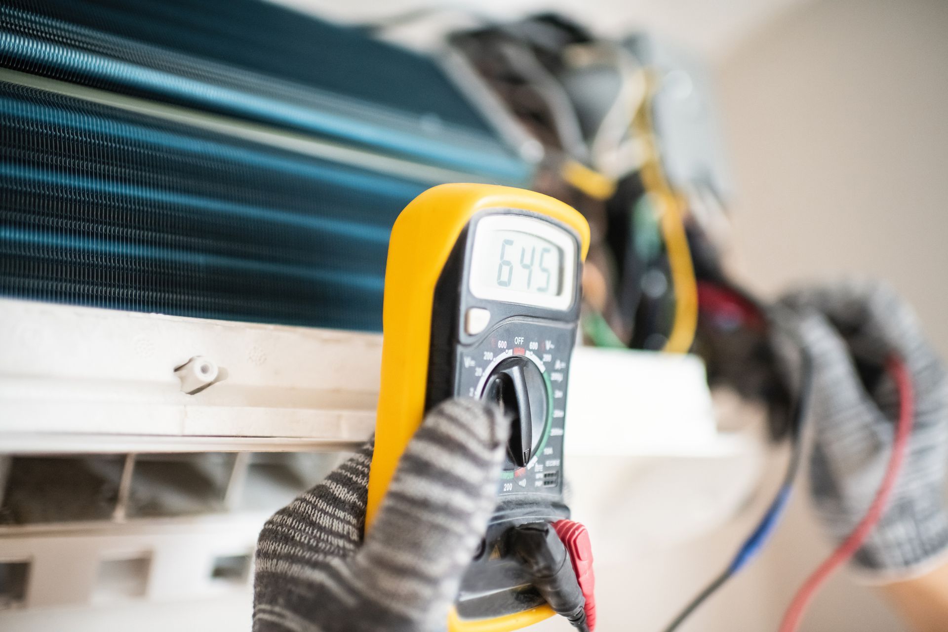 A person wearing gloves using a multimeter to test an air conditioning unit.