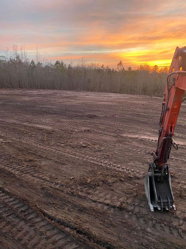 An excavator is sitting in the middle of a dirt field at sunset.