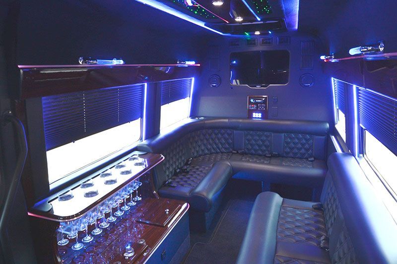 Inside of a black party bus with blue lighting, seating, bar with glasses, and TV.
