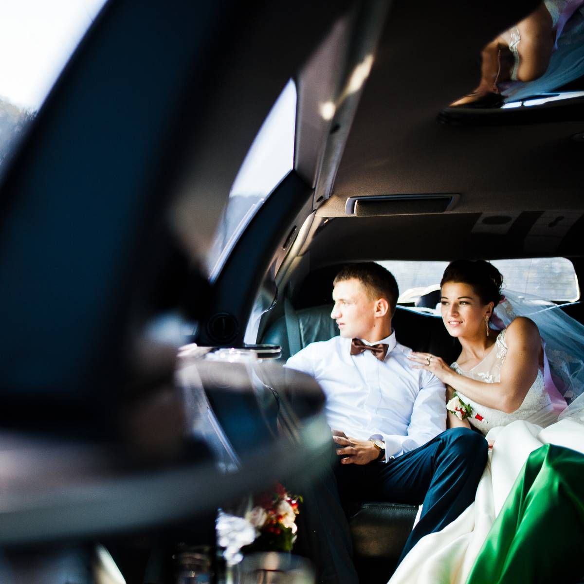 Bride and groom in a limousine, looking out the window. The groom wears a bow tie, and the bride is in a wedding dress.