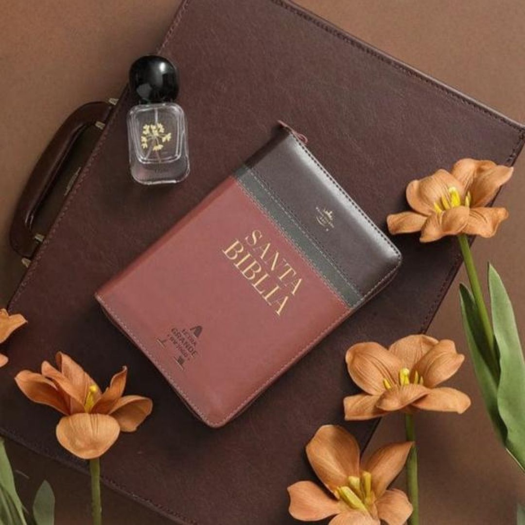 Biblia en español sobre cuaderno encuadernado en cuero con perfume y flores sobre superficie marrón.