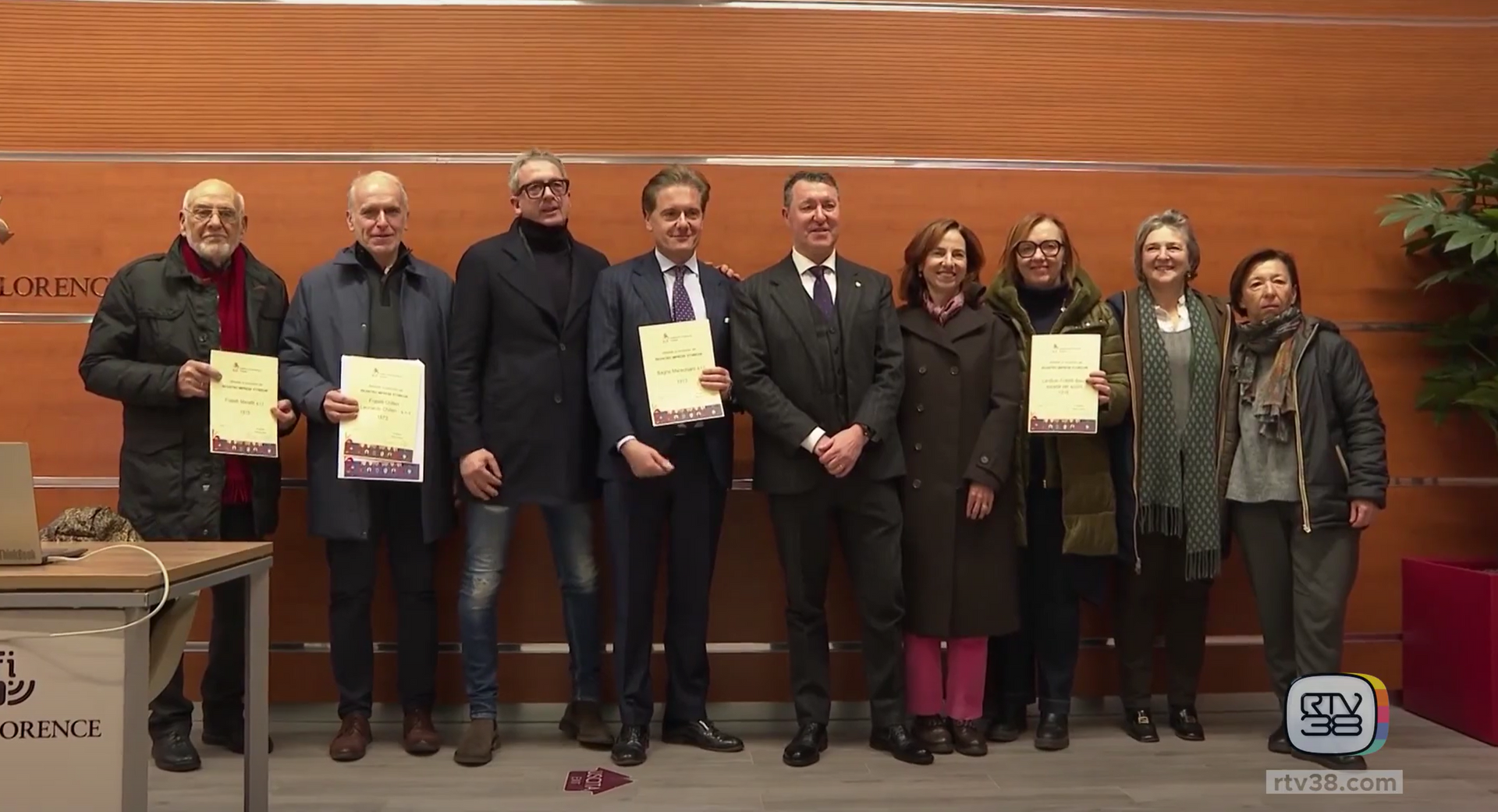 Gruppo di persone in posa in un interno vicino a un tavolo, con in mano dei certificati. Sfondo neutro.